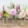Trio of small glass bud vases with blush roses, a pink dahlia, and delicate accent flowers.