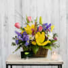 Basket arrangement of tulips, irises, and daisies with a candle beside it