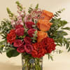 Birthday arrangement of red and orange roses, red carnations, and pink snapdragons in a glass vase.