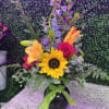 Tall mixed bouquet with sunflower, lilies, rose, and blue delphinium in a black vase with green bow.