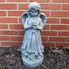 Blue-gray angel statue holding flowers in front of a brick wall