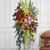 Tall standing floral spray with red roses, sunflowers, lilies, and blue accents beside a casket