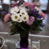 A purple vase with roses, daisies, carnations, and purple filler flowers for