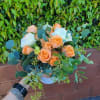 Low arrangement of peach roses and white blooms in a white bowl vase, held in front of a leafy hedge