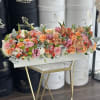 Low rectangular arrangement of pink, peach and orange flowers in a white box on a gold stand