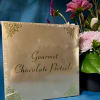 Gold box of gourmet chocolate pretzels beside a pink and purple flower arrangement on a teal background