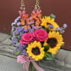 Bouquet of pink roses, sunflowers, and purple filler in a glass vase with a striped ribbon.