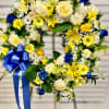 Standing wreath of white, yellow, and blue flowers with a large blue bow on an easel.