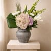 Asymmetrical arrangement of blush hydrangea, lavender roses, white lilies, and broad green leaves in a gray ceramic vase