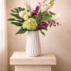 Tall white vase with white roses, lilies, green hydrangea and purple orchids with large tropical leaves.
