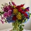 Tall mixed arrangement of red roses, yellow pincushion protea, blue hydrangeas, and spotted orchids in a clear glass vase