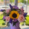 Round lavender hatbox with one bright sunflower, pink roses, carnations and purple