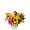 Short glass vase of bright orange lilies, pink roses, a sunflower, and mixed colorful blooms.