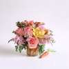 Short arrangement of pink roses, yellow orchid, and orange flowers in a carrot-filled glass vase with a carrot beside it