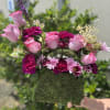Pink roses and magenta flowers in a moss-covered box with a gold 