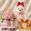 Pink rose bouquet in a scooter-shaped vase beside a teddy bear and chocolates
