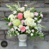 Tall arrangement of white and pink roses with pink carnations and green hydrangea in a white urn vase.