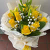 Hand-tied bouquet of yellow roses, lily buds, and white filler flowers wrapped in white paper.