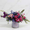 Asymmetrical arrangement of pink roses and purple and blue blossoms in an iridescent lavender vase