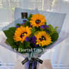 Hand-tied bouquet of yellow sunflowers with purple accents in blue wrap on a stand