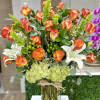 Tall arrangement of orange roses, white lilies, and green hydrangeas in a clear glass vase