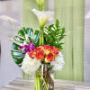 Tall arrangement of white calla lilies, orange roses, hydrangeas, and orchids in a glass vase with stones.