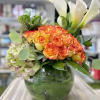Low round arrangement of orange roses, green hydrangea, and white calla lilies in a glass bowl vase