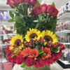 Tall arrangement of red roses, yellow sunflowers, and orange gerbera daisies in a round glass vase.