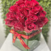 Dense bouquet of red roses arranged in a square glass vase wrapped with a red ribbon.