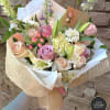 Hand-tied bouquet of pastel roses, lilies, and mixed blooms wrapped in white and burlap-style paper
