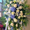 Tall standing spray of white roses, lilies, and blue hydrangeas with palm fronds on an easel.