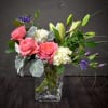 Arrangement of pink roses, white hydrangeas, and lilies in a clear glass cube vase with purple accents