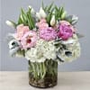 Pink peonies, pale roses, white tulips, and hydrangeas arranged in a clear glass cylinder vase with stones