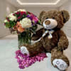 Bouquet of mixed flowers beside a teddy bear with a ribbon and pink heart confetti