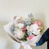 Hand-tied bouquet of pastel pink, white, and blue flowers wrapped in paper