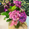 Pink roses and purple flowers arranged in a glass vase with a pink ribbon.