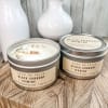 Two lidded tin candles labeled Black Currant + Jasmine on a wood block with white vases behind