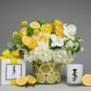 Yellow and white floral arrangement in a lemon-decorated vase with a gift box and candle
