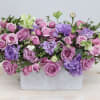Low rectangular arrangement of pink and lavender roses with purple blooms in a white container