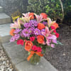 Mixed bouquet of pink lilies, orange roses, and purple daisies in a glass vase