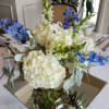 Centerpiece of white hydrangeas, snapdragons, and blue delphinium in a clear vase on a mirror.