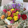 Handheld bouquet of multicolored roses, dahlias, and purple blooms wrapped in pink paper