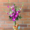 Tall vase of purple and white orchids with white filler flowers and a small teddy bear hugging the vase