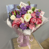 Hand-tied bouquet of pink roses, purple flowers, and white blooms wrapped in pink paper with a sheer ribbon.