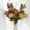 Tropical flower arrangement in a tall clear vase with purple orchids and yellow pincushion blooms