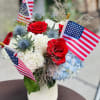 Red roses and blue hydrangeas in a white vase with small American flags