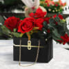Red roses arranged in a black handbag-style box with gold chain handles.