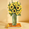 Tall bouquet of yellow sunflowers in a clear glass vase on a colorful book against a yellow background