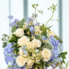 Compact arrangement of cream spray roses, lavender hydrangeas, and small daisies in a round vase.