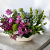 Low white ceramic arrangement with purple roses, orchids, and white tulips in an asymmetrical design